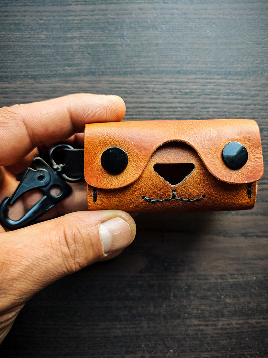 Doggy Duty leather waste bag dispenser featuring a dog face design created with snap eyes, a cutout nose, stitched mouth, and blue waste bag dispensing opening.
