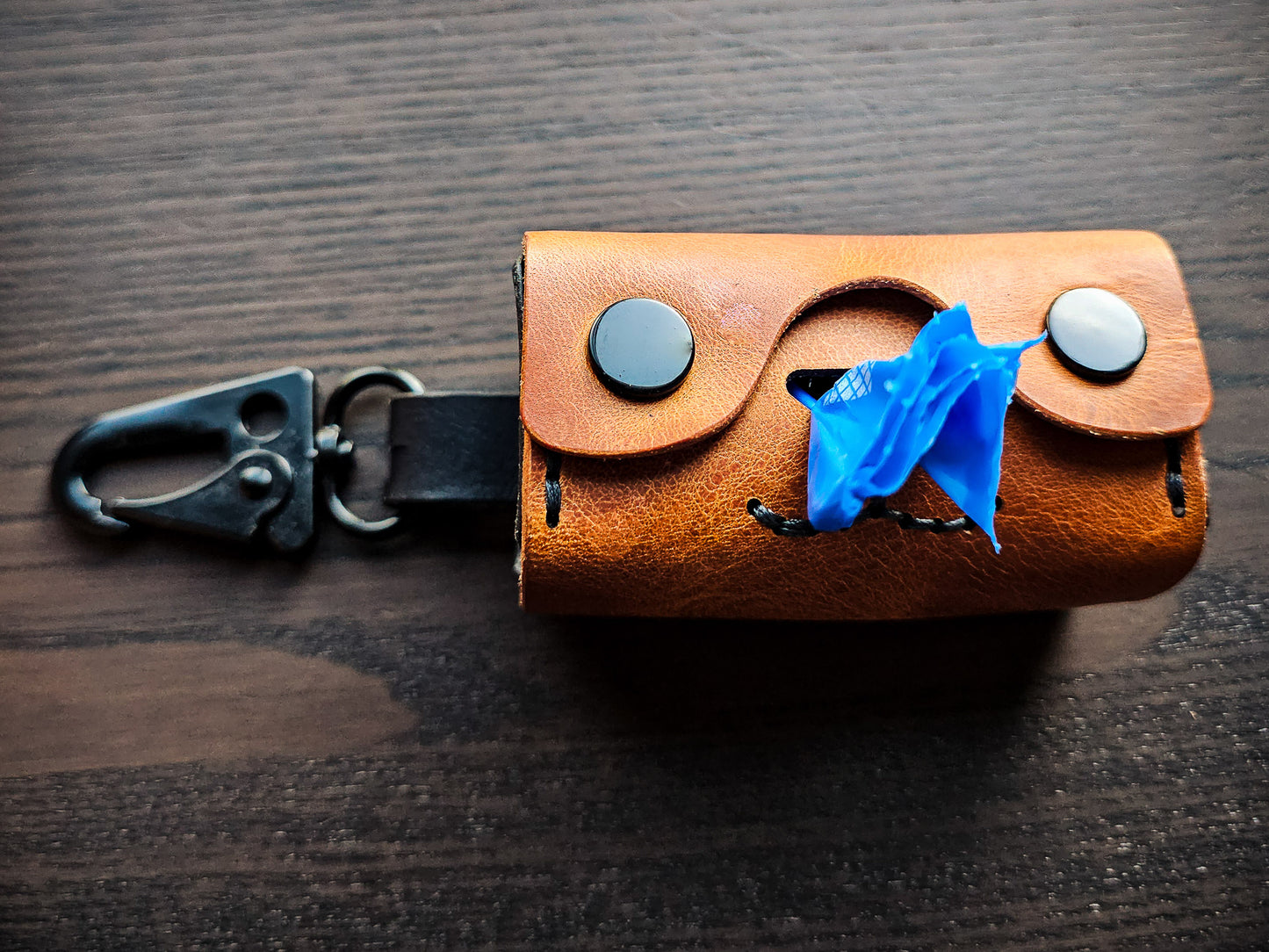 Doggy Duty brown leather waste bag dispenser resting on a tabletop with black swivel clip and blue waste bag visible through the dispensing opening.