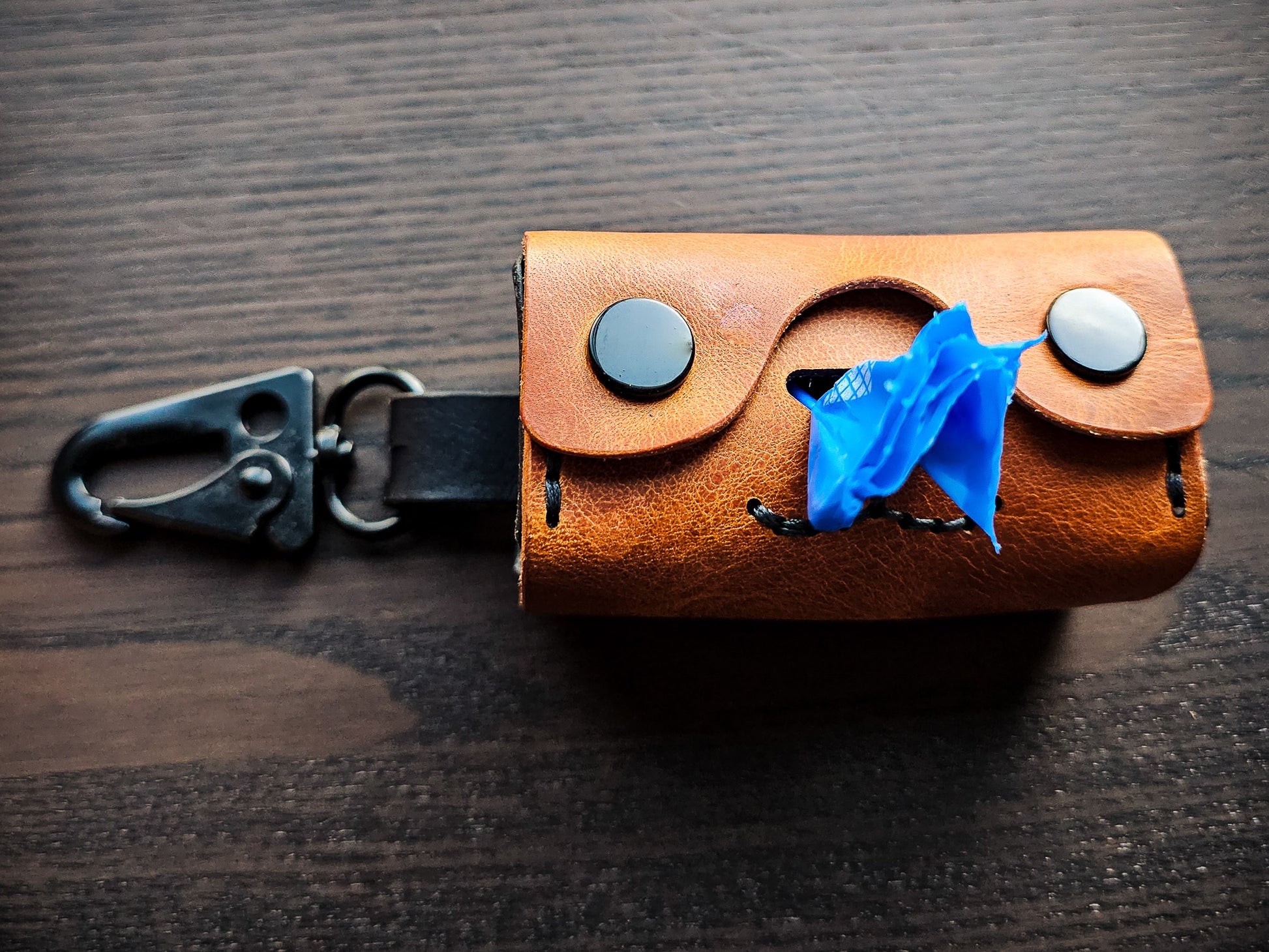 Doggy Duty brown leather waste bag dispenser resting on a tabletop with black swivel clip and blue waste bag visible through the dispensing opening.