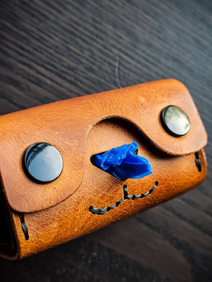 Close-up of the Doggy Duty leather waste bag dispenser showing snap eyes, cutout nose, stitched mouth, and waste bag dispensing opening.