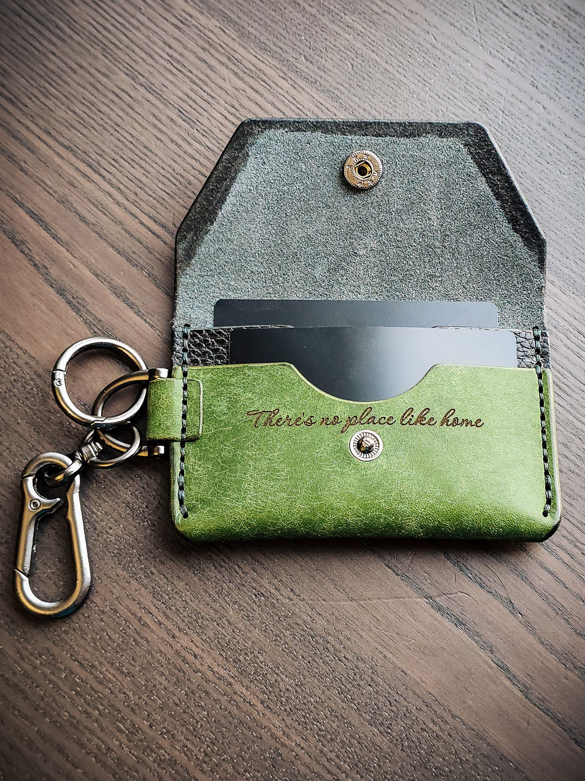 Green leather cardholder and keychain engraved with the phrase "There's no place like home," representing the connection between personal objects, memory, and lived experience.