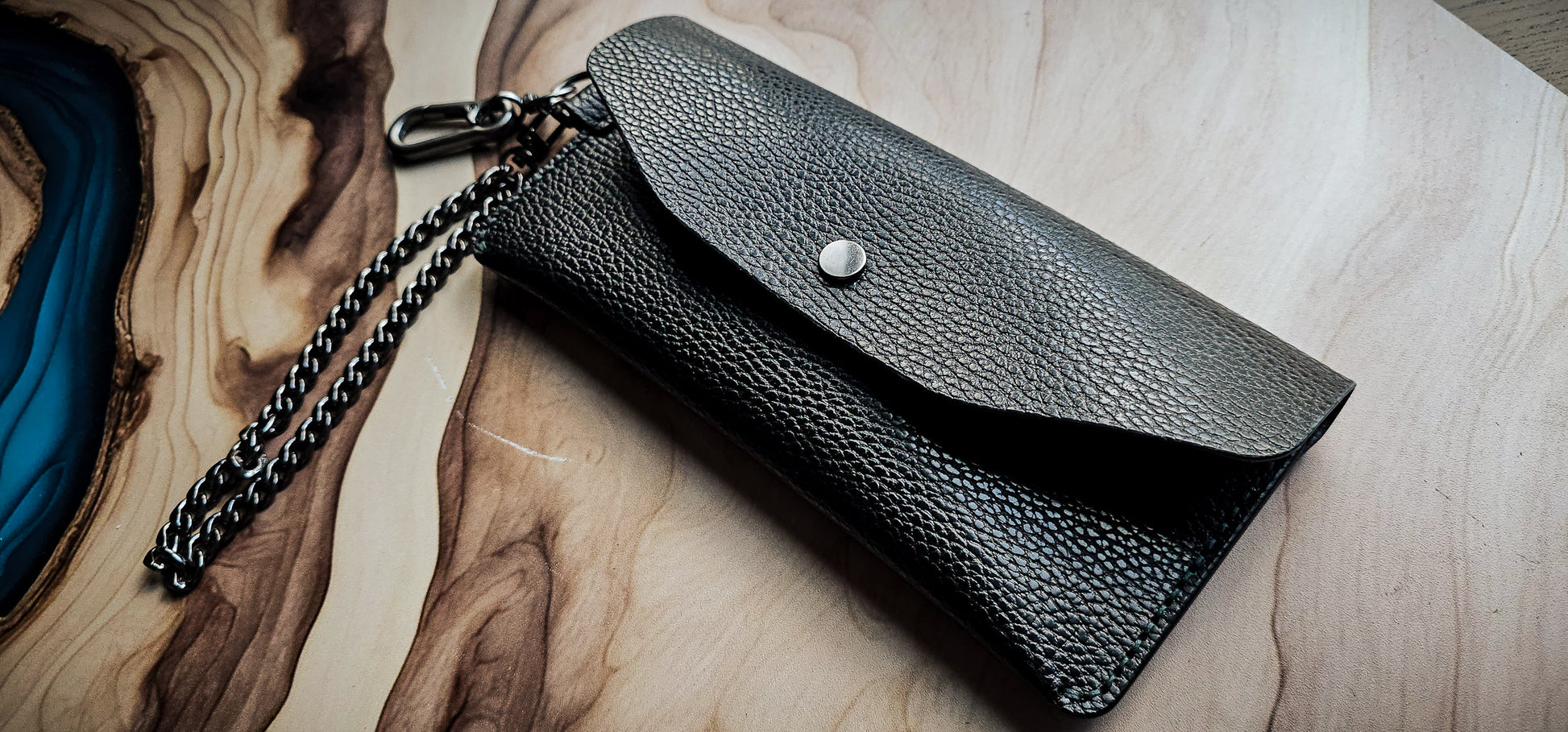 Delilah Clutch handmade from black pebbled leather with silver snap closure and chain strap, photographed on a wood and resin surface.