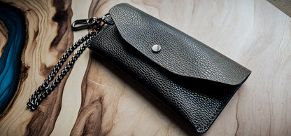 Delilah Clutch handmade from black pebbled leather with silver snap closure and chain strap, photographed on a wood and resin surface.