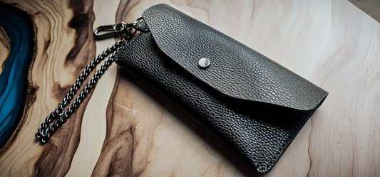 Delilah Clutch handmade from black pebbled leather with silver snap closure and chain strap, photographed on a wood and resin surface.